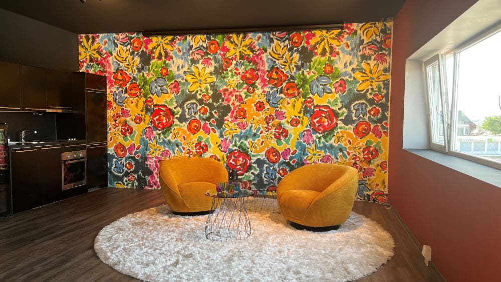 Designertapete MARNI von Londonart im Hintergrund und 2 orangen Sesseln und 1 Glastisch auf rundem Teppich im Vordergrund - links davon eine Küchenzeile